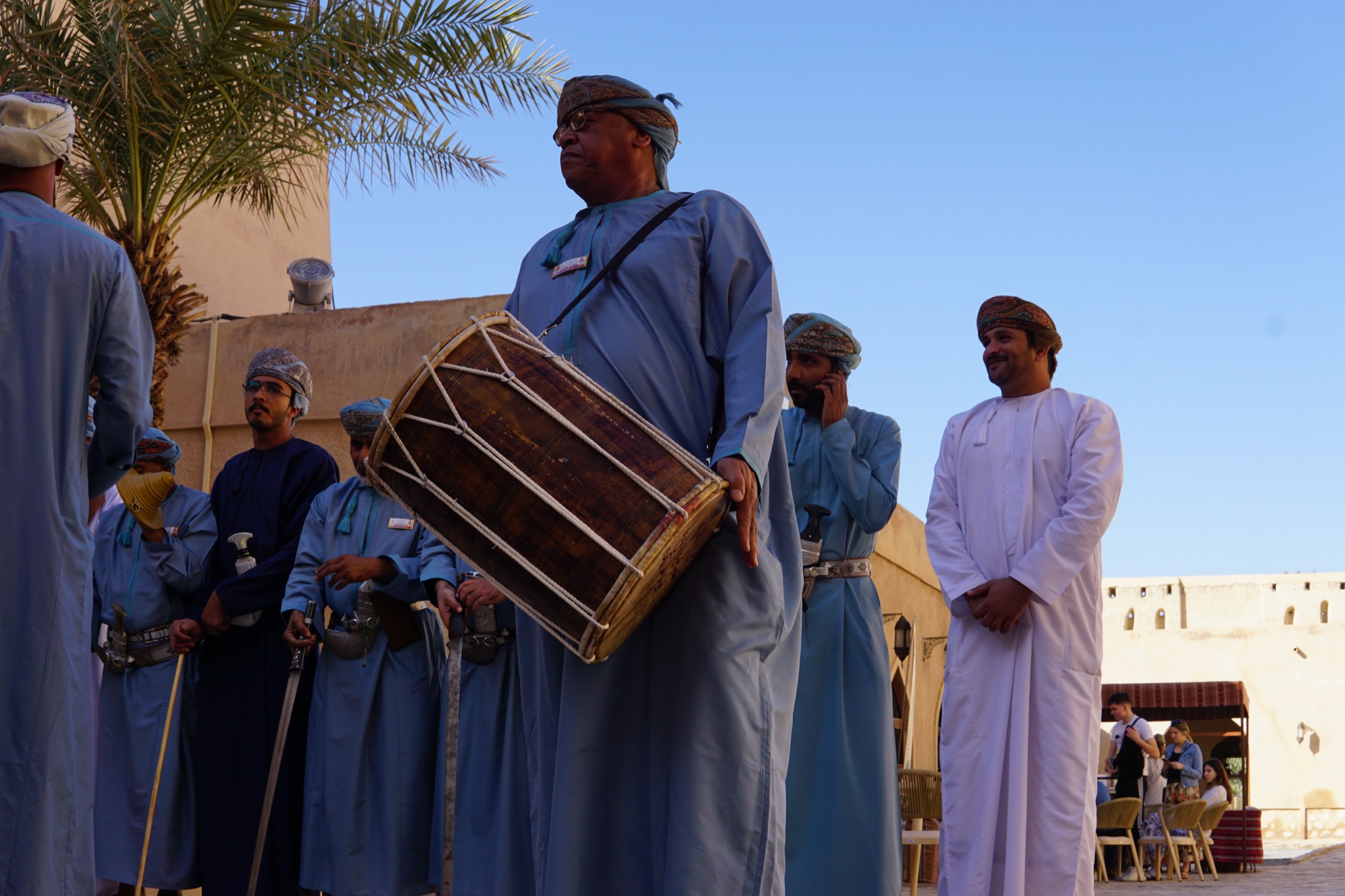 A drummer at a fort in Oman holding a rope-tensioned drum, men in blue dishdashas beside him